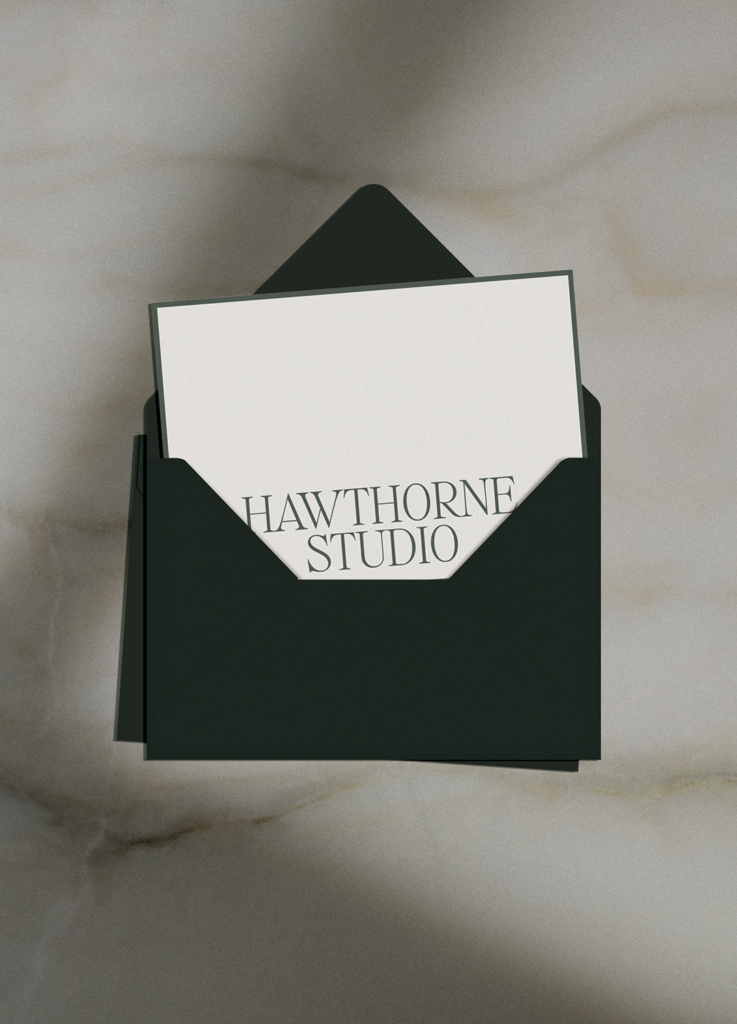 A white card with the text HAWTHORNE STUDIO in elegant capital letters is partially pulled out of a dark green envelope, placed on a light marble surface.