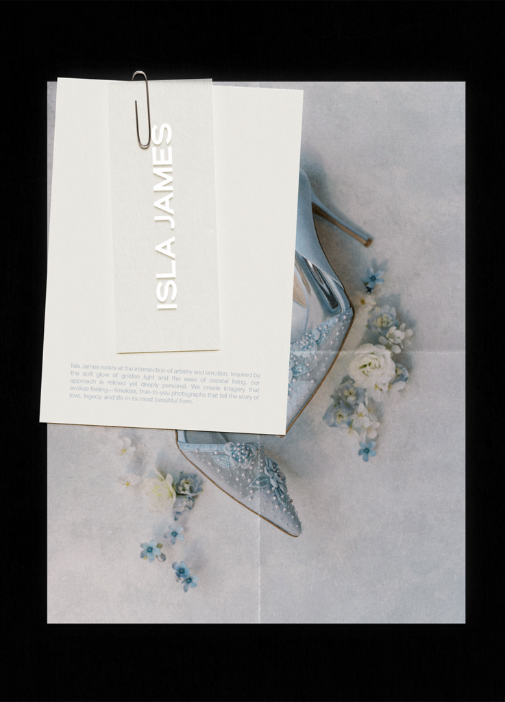 A folded card with the name Isla James clipped on top, covering part of a photo showing elegant, embellished silver high-heeled shoes surrounded by light blue and white flowers.