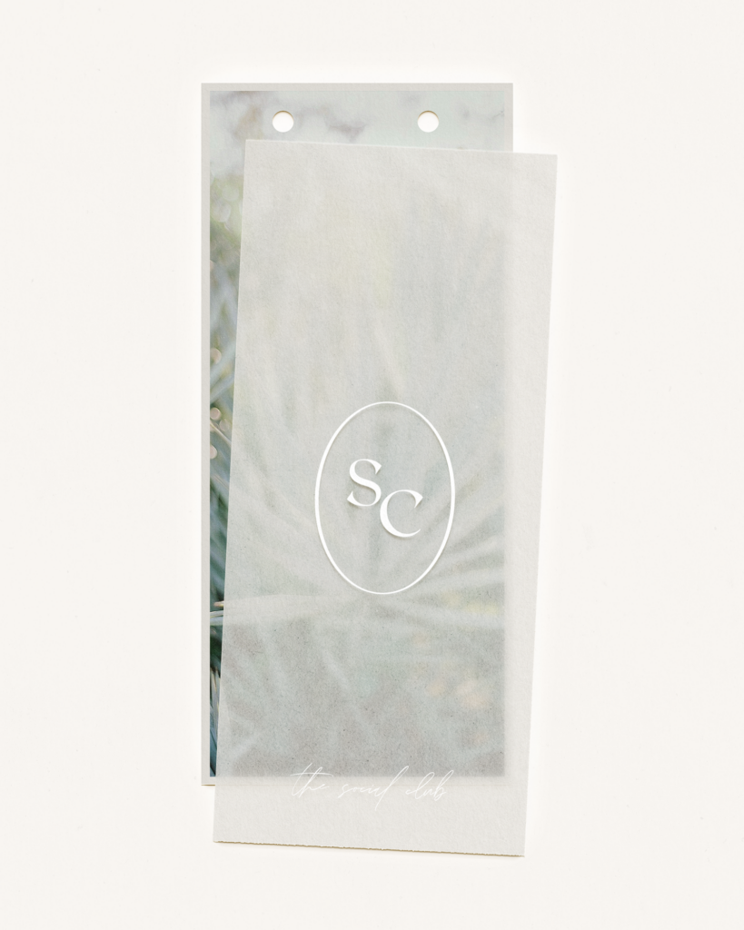 A translucent sheet with an SC monogram inside an oval is layered over a blurred image of green foliage. Two holes are punched at the top of both sheets. The words “the social club” are faintly visible at the bottom.