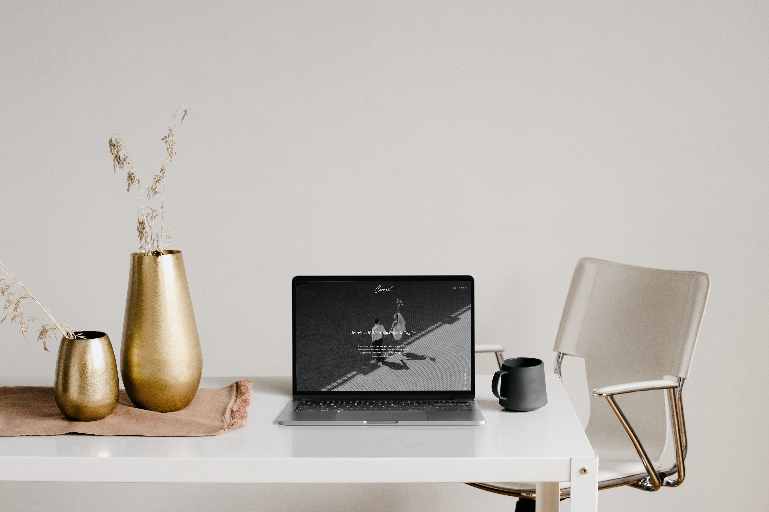 A minimalist workspace with a white desk, beige chair, gold vases with dried grass, a black coffee mug, and an open laptop displaying a grayscale image on the screen.