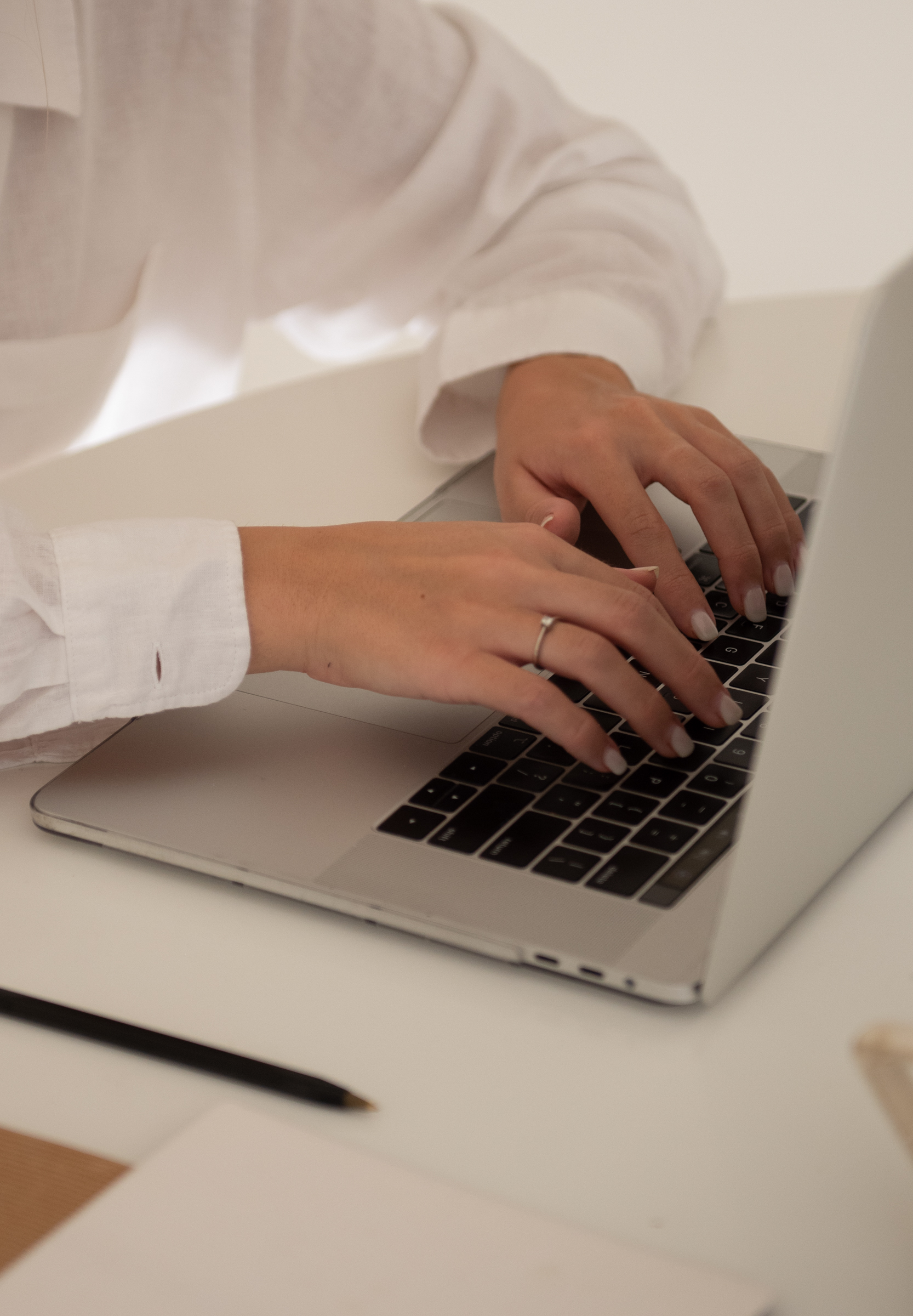 A person wearing a white shirt is typing on a laptop keyboard, perhaps researching Showit vs Squarespace vs Shopify. Only their hands and part of their torso are visible, with a pencil and some papers on the white desk nearby.