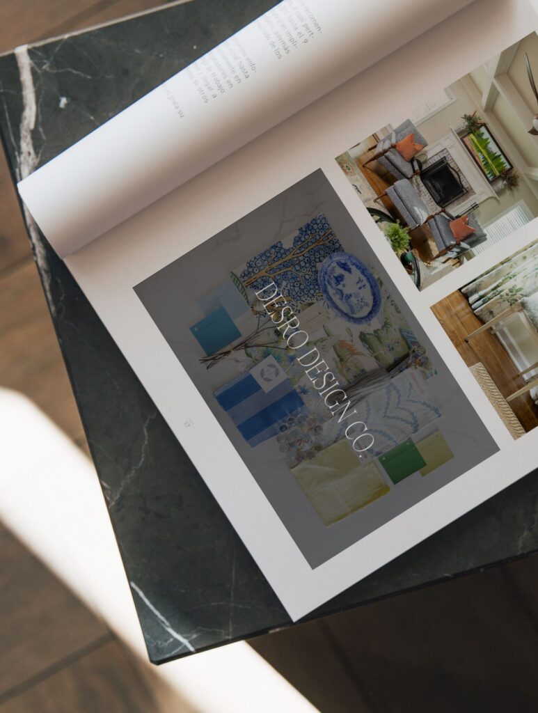 An open magazine lies on a dark marble table, displaying pages with interior design photos, color swatches, and the text DESIGN DESIGN CO. across one of the images. Sunlight partially illuminates the scene.