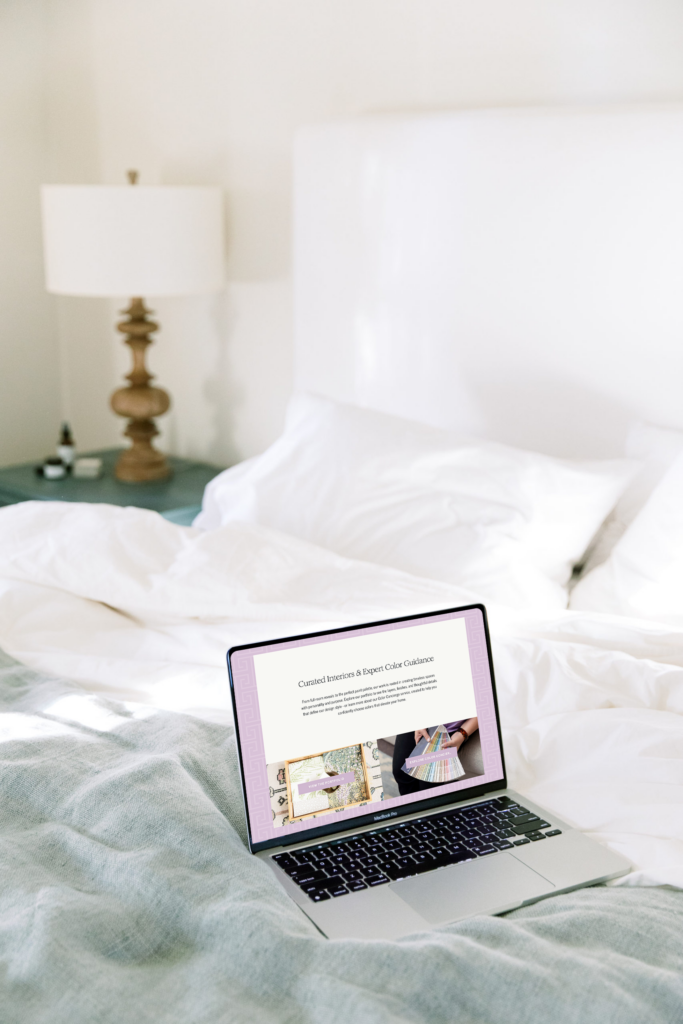 A laptop displaying a colorful website rests on a neatly made bed with white bedding; a bedside table with a lamp and small bottles is in the background.
