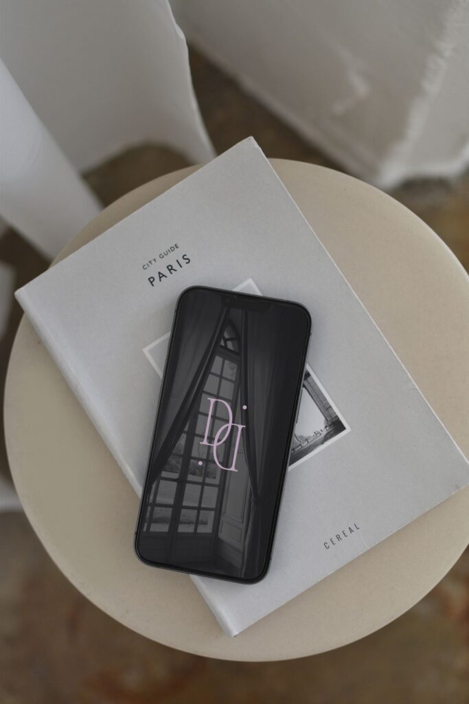A smartphone with a minimalistic logo on its screen rests on a white Paris city guide book, placed on a round beige table near a window.