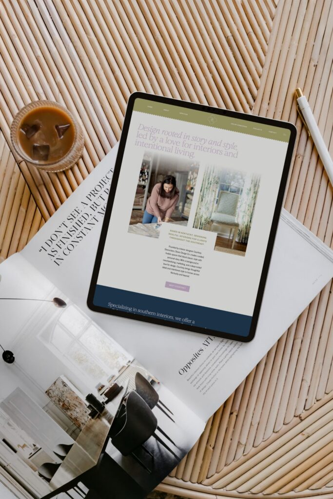 A tablet showing an interior design website rests on a wicker table beside an iced coffee and an open magazine featuring modern home interiors.