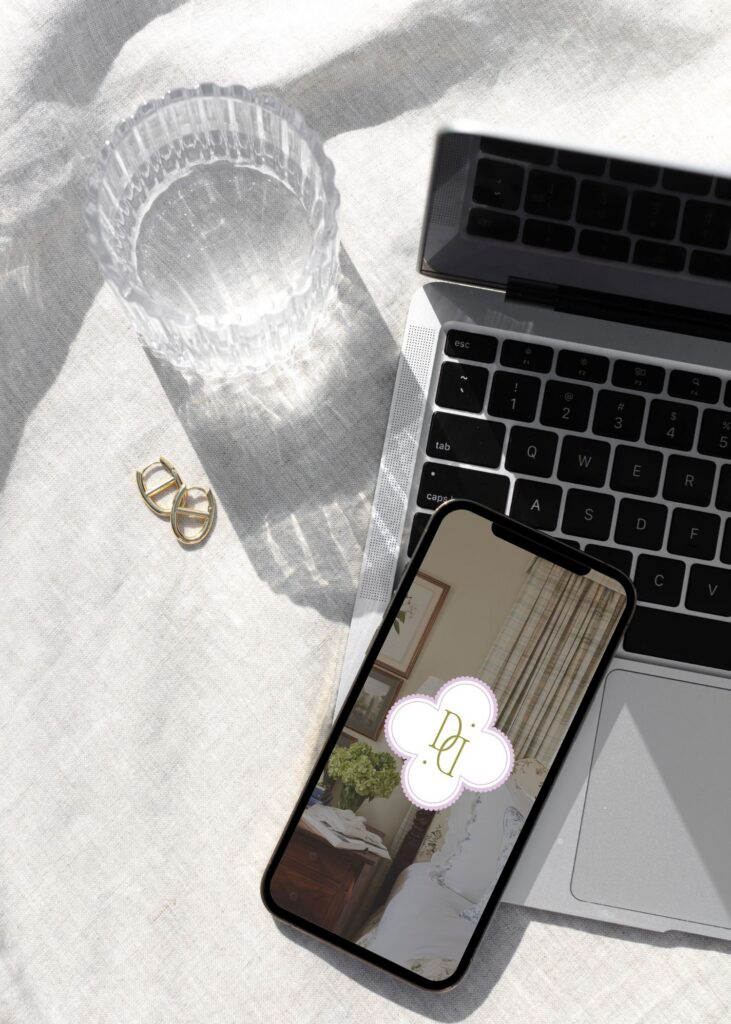 A smartphone with a decorative logo on its screen rests on a laptop keyboard. Nearby are a pair of gold earrings and a clear, empty glass, all on a light textured fabric surface in bright sunlight.