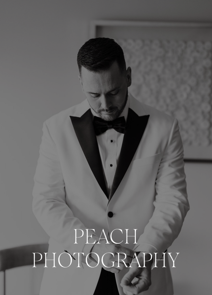 A man in a white tuxedo with a black bow tie stands indoors, adjusting his cufflinks. The image is black and white and features the text PEACH PHOTOGRAPHY at the bottom.