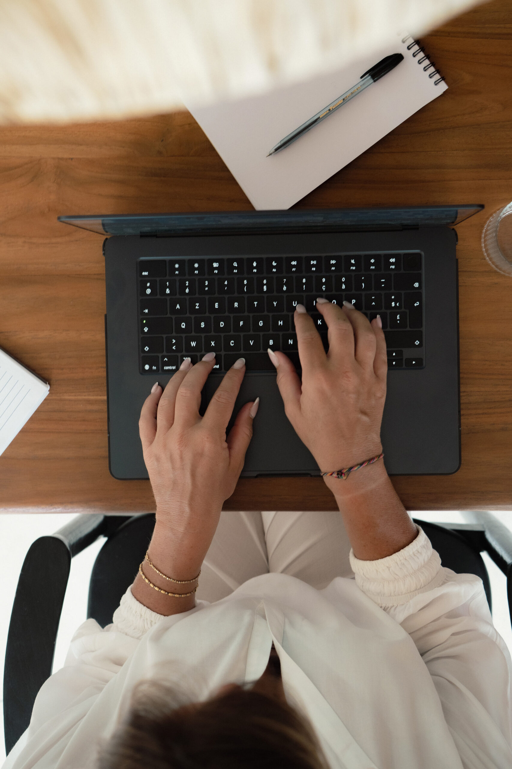 A person wearing a white shirt types on a black laptop at a wooden desk, with a notepad, pen, and glass of water nearby.