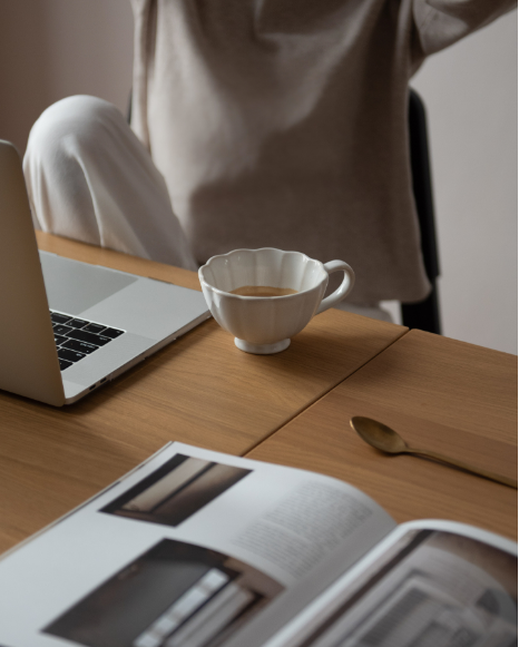 A person sits at a desk with a laptop, a cup of coffee, an open magazine, and a gold spoon on a wooden surface. The scene appears calm and cozy.