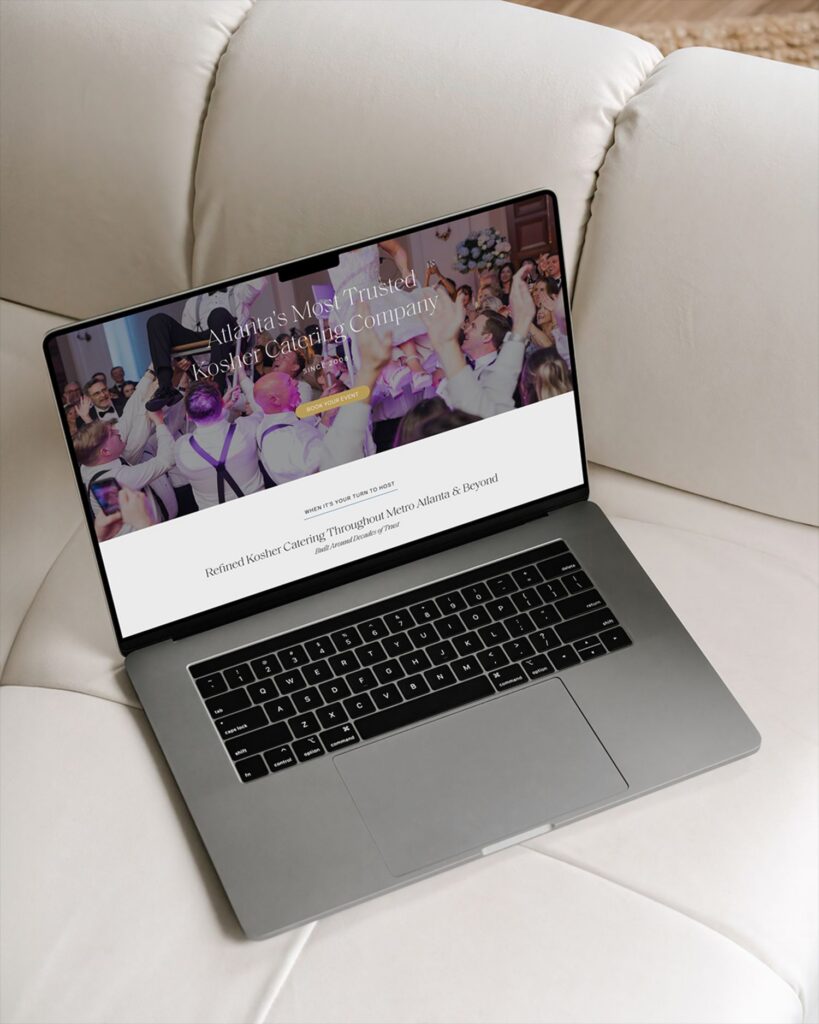 A laptop sitting on a white couch displays a catering company website featuring a photo of people celebrating at a wedding.